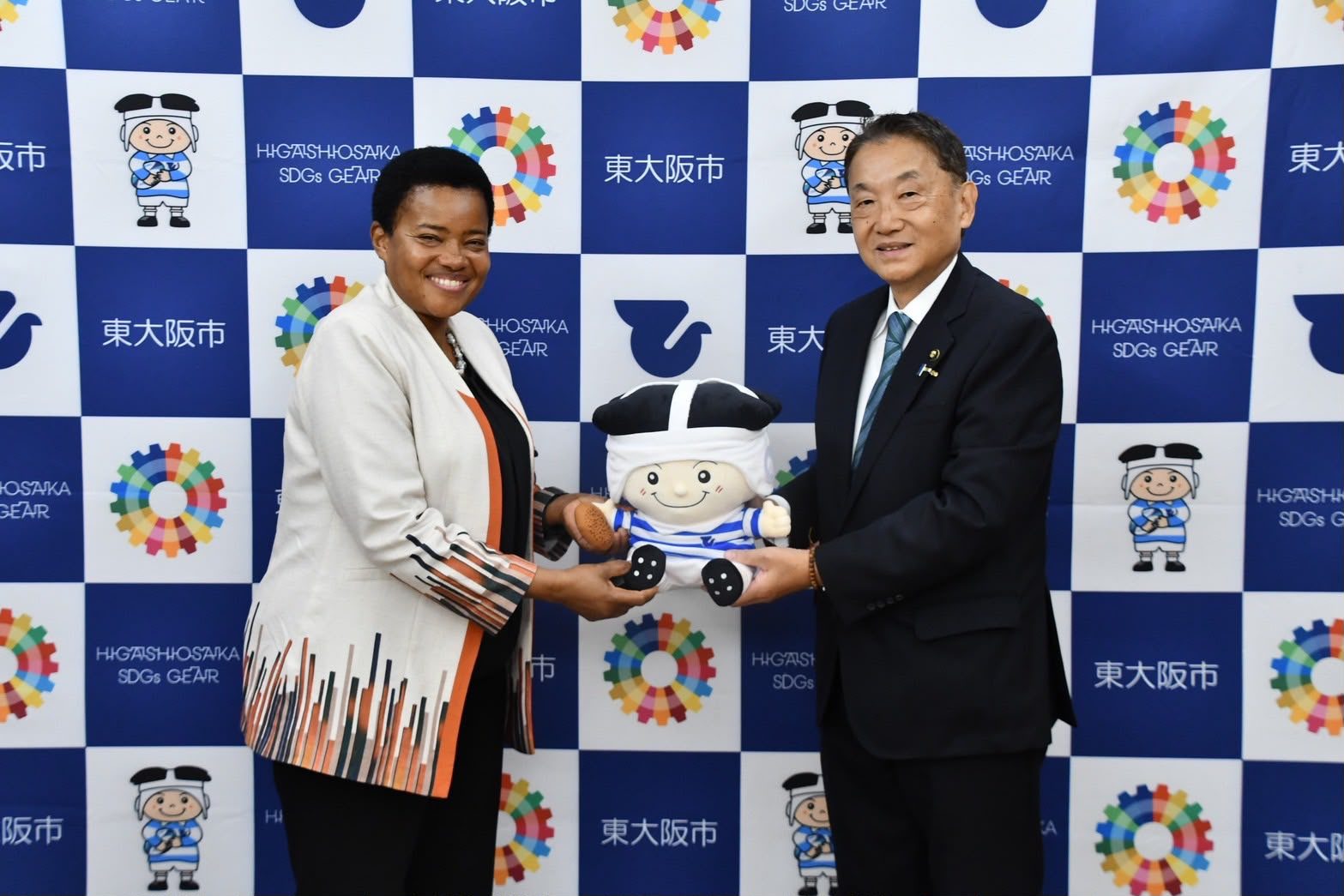 A representative and the Mayor of Higashiosaka City exchanging a gift in front of a Higashiosaka SDGs backdrop