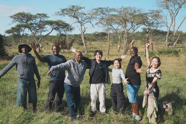 A mixed Ugandan and Japanese group on a safari visit, with giraffes in the grassland behind them